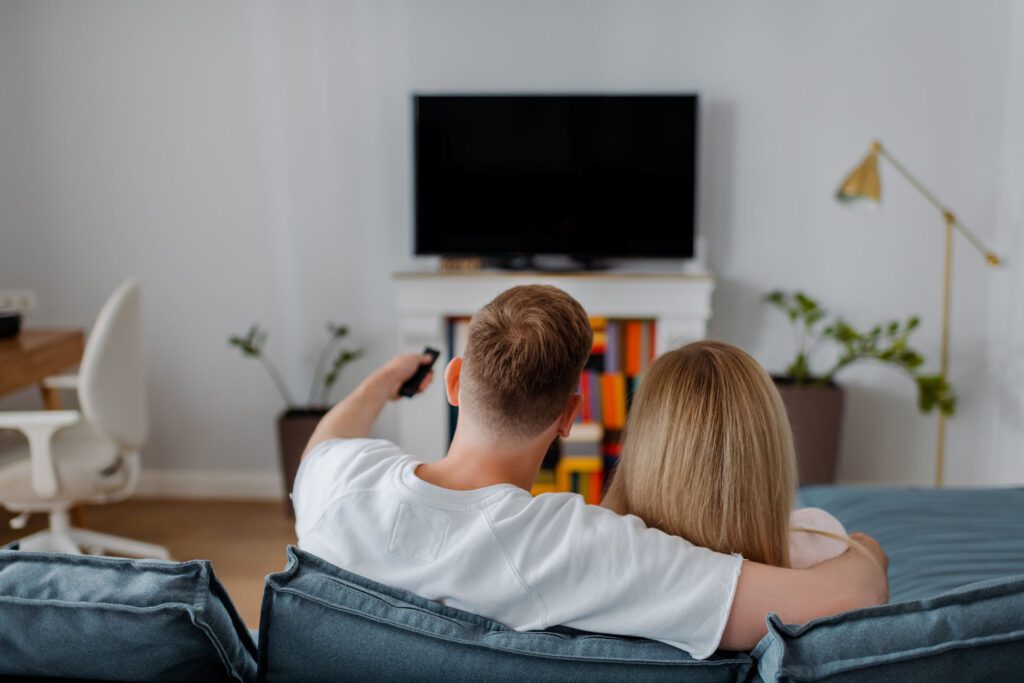 Couple on a couch sitting next to a TV, lamp, plants, and a desk with a rolling chair.