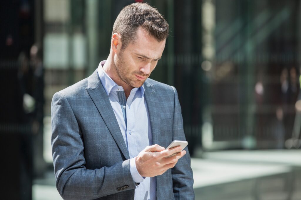 A businessman in a suit scrolls through a mobile phone in a city setting.