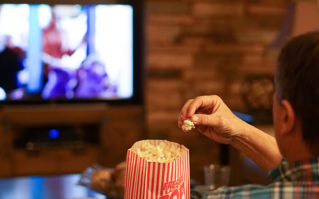 A man sits on a couch eating popcorn and watching a movie on television in his home.