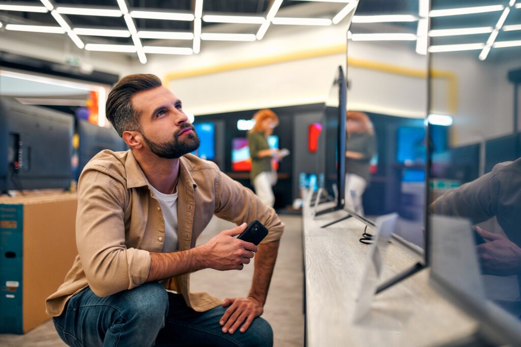 A man crouches to inspect a TV in a store full of TVs.