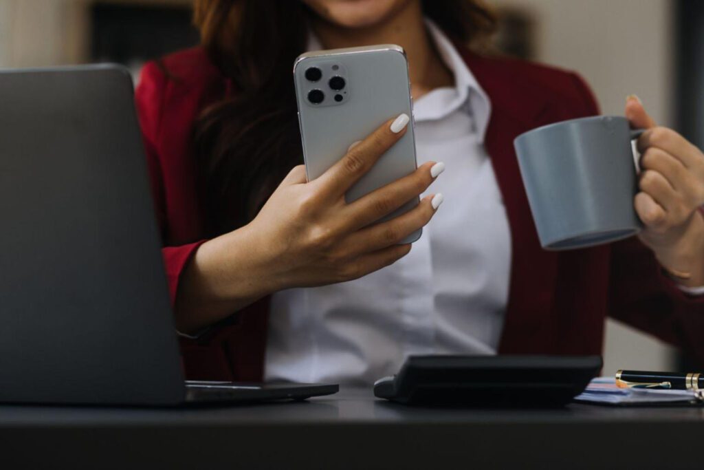 Woman using a cell phone and holding a mug next to a laptop, calculator, and a pen.