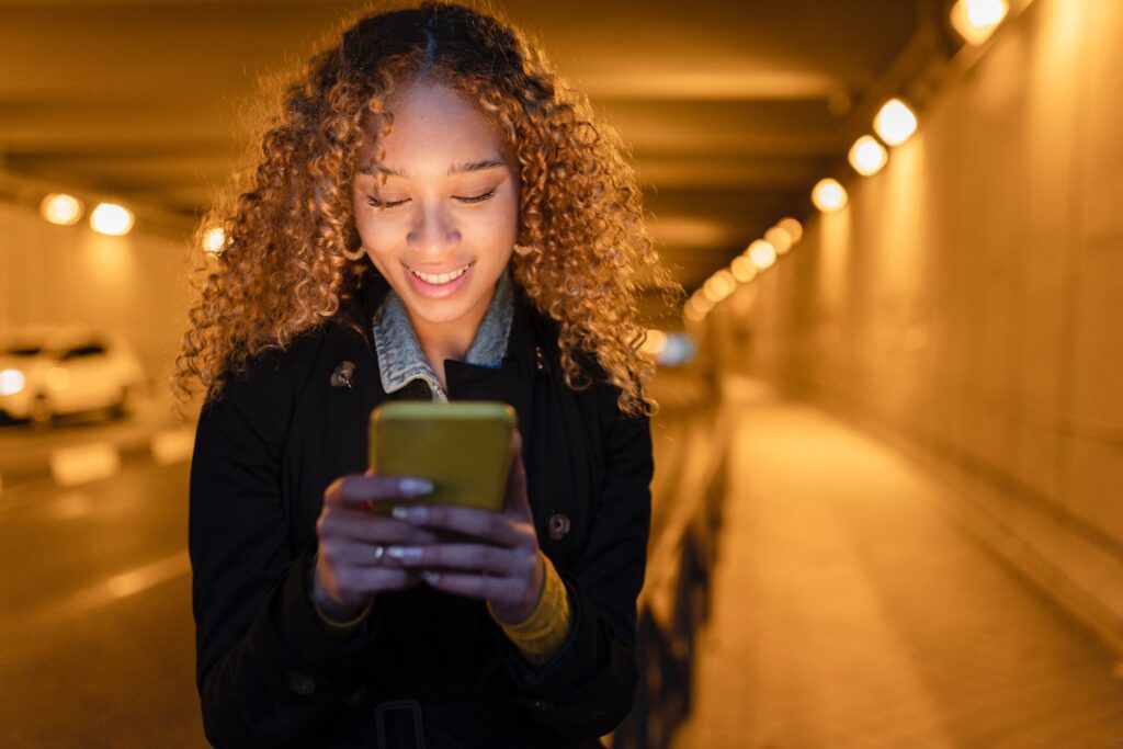 A smiling young woman walks through a lit tunnel at night while looking at her phone. 