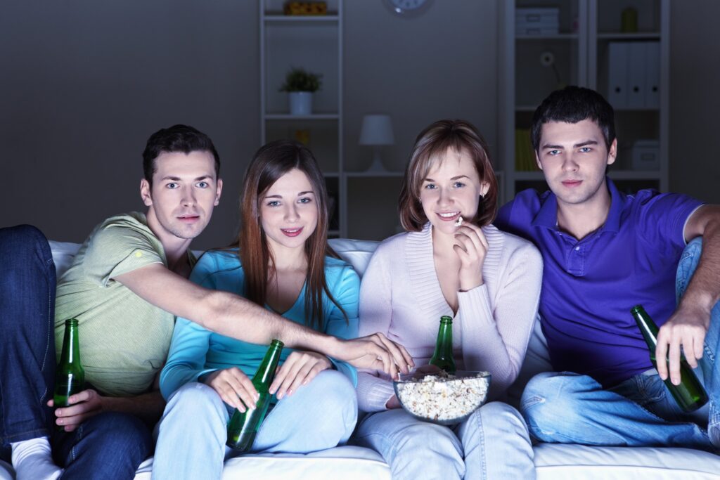 A group of friends eats from a bowl of popcorn as they watch TV. They are sitting in the dark, lit up by the screen.